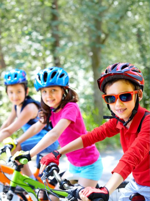 Tres niños con cascos montan en bicicleta al aire libre en un día soleado y sonríen a la cámara. El fondo está lleno de árboles verdes.