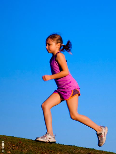 Joven con un conjunto deportivo rosa corriendo al aire libre en una colina con un cielo azul claro de fondo.