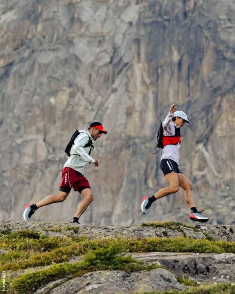 Dos personas caminan por un sendero rocoso frente a un escarpado acantilado de montaña, llevan sombreros, mochilas y ropa deportiva.