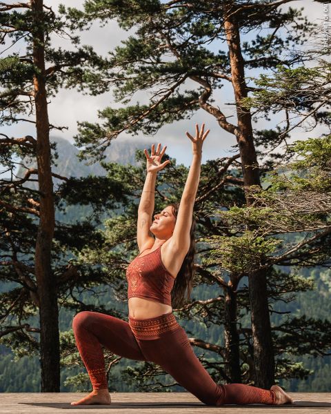 Una mujer con ropa deportiva roja practica yoga al aire libre en una terraza de madera, rodeada de altos árboles y montañas, y estira los brazos por encima de la cabeza en una postura de estocada.