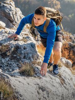 Un joven con una sudadera azul y una mochila escala una ladera rocosa de montaña, rodeado de follaje otoñal.