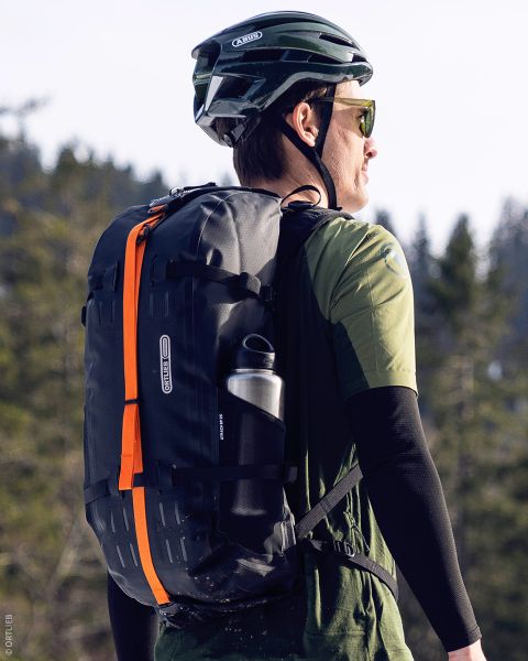 Una persona que lleva un casco verde y gafas de sol, una mochila negra con una franja naranja y una botella de agua, se encuentra al aire libre en una zona boscosa.