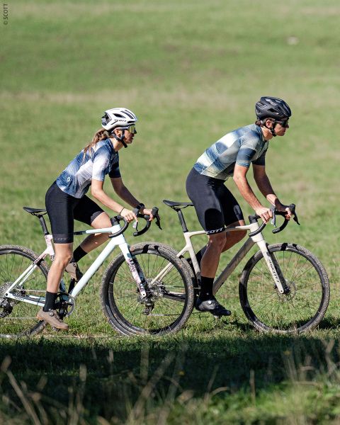 Dos ciclistas con cascos y ropa de ciclismo montan en bicicletas blancas uno al lado del otro por un camino de hierba, con un prado verde al fondo.
