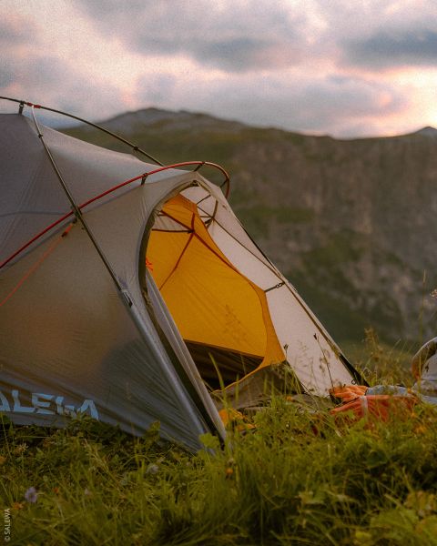 Una tienda de campaña gris y amarilla se encuentra en un prado con montañas al fondo bajo un cielo nublado al atardecer.