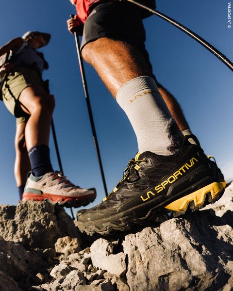 Dos excursionistas están de pie con bastones de trekking en un terreno rocoso; el foco está en uno de ellos, que lleva zapatillas La Sportiva negras y calcetines blancos bajo un cielo azul despejado.
