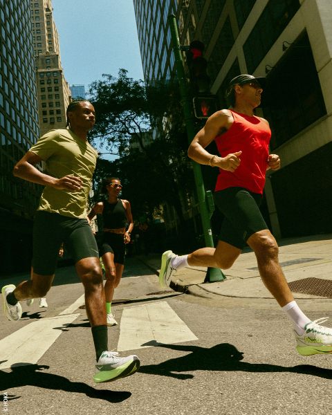 Tres personas corren con ropa deportiva y zapatillas por una calle de la ciudad, con edificios altos y luz solar al fondo.