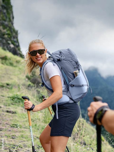 Una mujer con gafas de sol camina por un sendero de montaña cubierto de hierba con bastones de trekking y una mochila gris, sonriendo a la cámara.