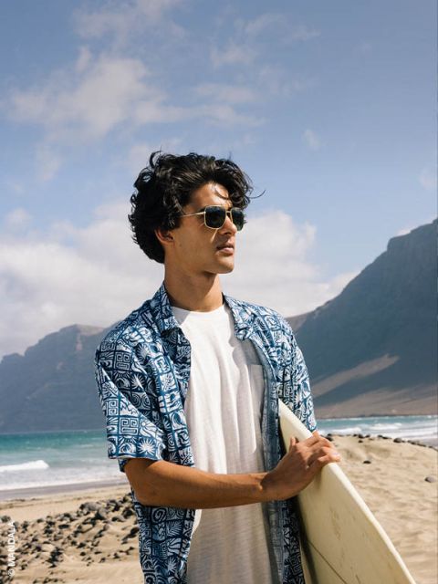 Hombre con gafas de sol y camisa azul estampada sostiene una tabla de surf y está de pie en una playa de arena con montañas y el mar de fondo.