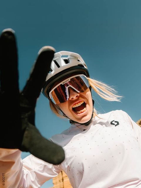 Un ciclista sonriente con casco, gafas de sol y guantes se estira hacia la cámara frente a un cielo azul radiante.