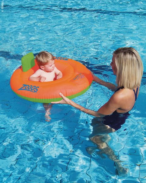 Una mujer sostiene a un bebé en brazos, que está sentado en un flotador naranja y verde en una piscina.