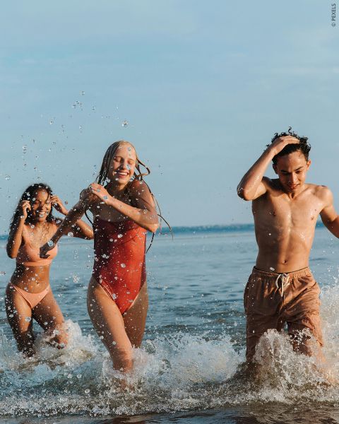Tres jóvenes adultos en traje de baño ríen y chapotean mientras caminan por aguas marinas poco profundas en un día soleado.