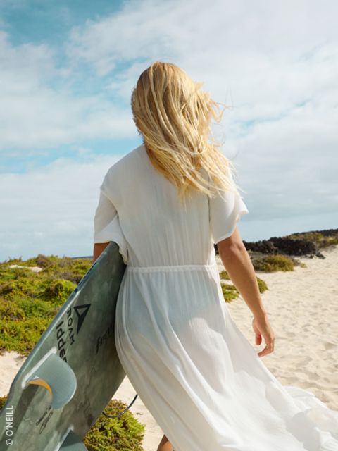 Una mujer con un vestido blanco camina con una tabla de surf en la mano por una playa de arena, su cabello rubio ondea al viento.