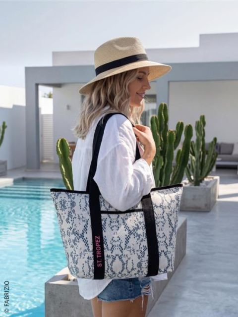 Una mujer con sombrero de paja y camisa blanca está de pie junto a una piscina y sostiene un bolso de mano grande y estampado con la inscripción "St. Tropez" en la mano, con cactus de fondo.