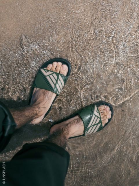 Una persona con sandalias verdes está de pie en el agua poco profunda de una playa de arena, con los pantalones ligeramente remangados.