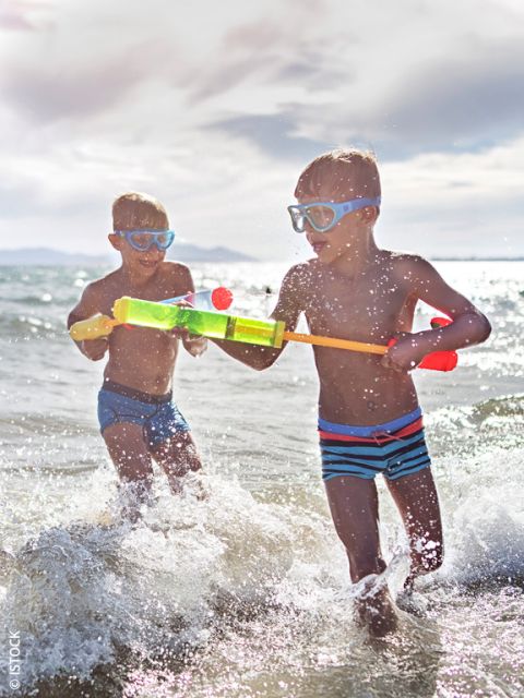 Dos niños con gafas de natación juegan con una pistola de agua en el mar y chapotean en las olas bajo un cielo brillante y soleado.