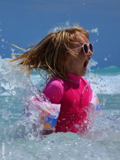 Una chica con gafas de sol rosas y traje de baño chapotea en el mar y sonríe emocionada mientras el agua salpica a su alrededor en un día soleado.