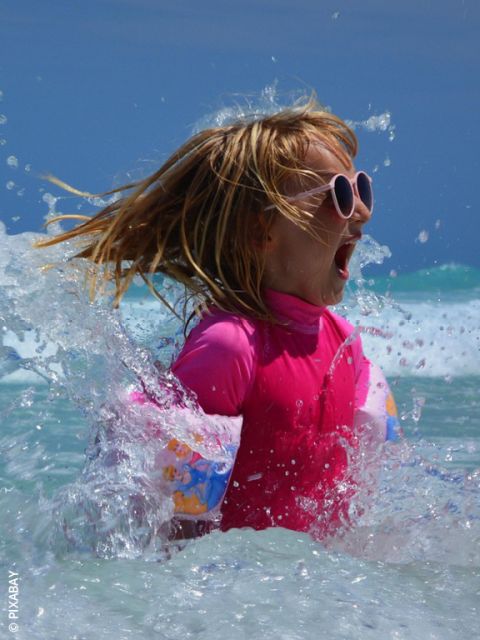 Una chica con gafas de sol rosas y traje de baño chapotea en el mar y sonríe emocionada mientras el agua salpica a su alrededor en un día soleado.