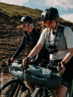 Dos mujeres con cascos y gafas de sol montan sonriendo en bicicletas al aire libre, a las que se les han sujetado bolsas. Al fondo se ve una colina cubierta de hierba.