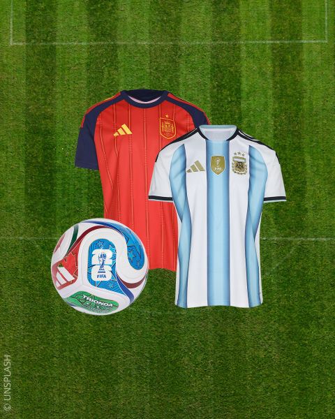 Dos camisetas de fútbol - una roja y otra blanca con rayas azules - junto a un balón de fútbol colorido en un campo de césped verde.