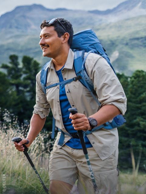 Un hombre que camina por la naturaleza con bastones de senderismo, lleva una mochila, una camisa beige y pantalones cortos, y sonríe con un fondo de árboles verdes y montañas.