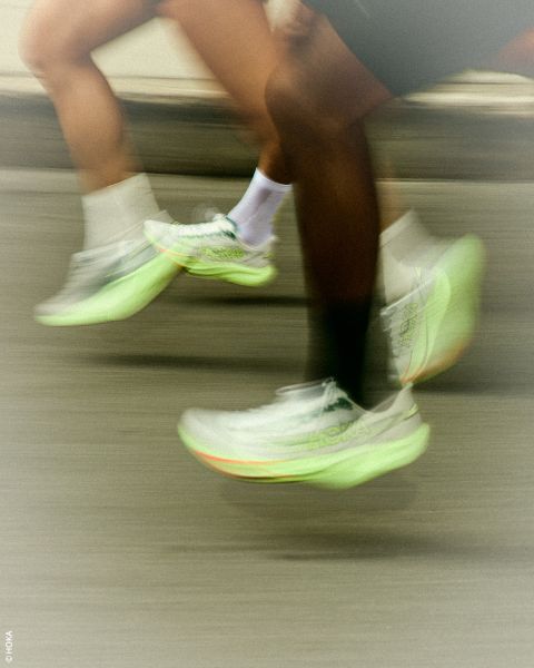 Dos personas corriendo con zapatillas HOKA de color verde neón; el desenfoque de movimiento subraya su rápido movimiento en una calle.