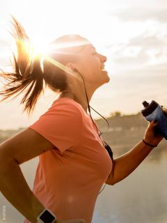 Una mujer con una camiseta rosa, que corre al aire libre al amanecer, sonríe, lleva auriculares y sostiene una botella de agua en la mano, mientras el sol brilla detrás de ella.
