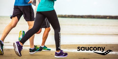 Tres personas corren con ropa deportiva a lo largo de una playa de arena; en la esquina inferior derecha se ve el logo de Saucony.