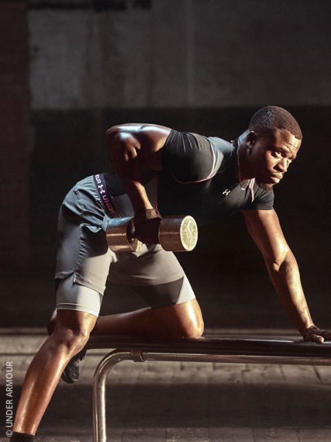 Un hombre en ropa deportiva levanta una mancuerna mientras se apoya en un banco; entrena concentrado en un gimnasio tenuemente iluminado.