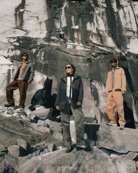 Tres personas con ropa de exterior de pie sobre terreno rocoso, con una gran pared de piedra detrás de ellas, mirando a lo lejos bajo un sol brillante.
