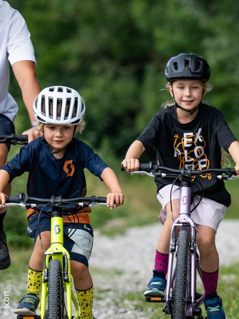 Dos niños pequeños con cascos montan en bicicleta afuera en un camino de grava, mientras un adulto está cerca. En el fondo borroso se ve vegetación.