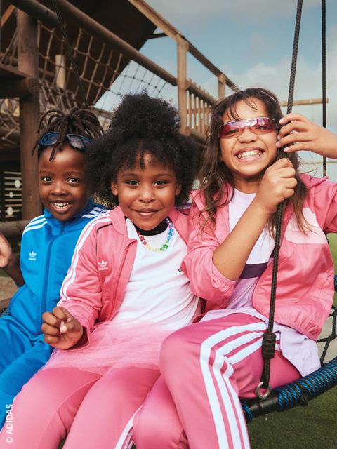 Tres niños sonrientes con atuendos de Adidas están sentados afuera en un columpio - dos de ellos con chándales rosas y uno con uno azul - y disfrutando del día soleado.