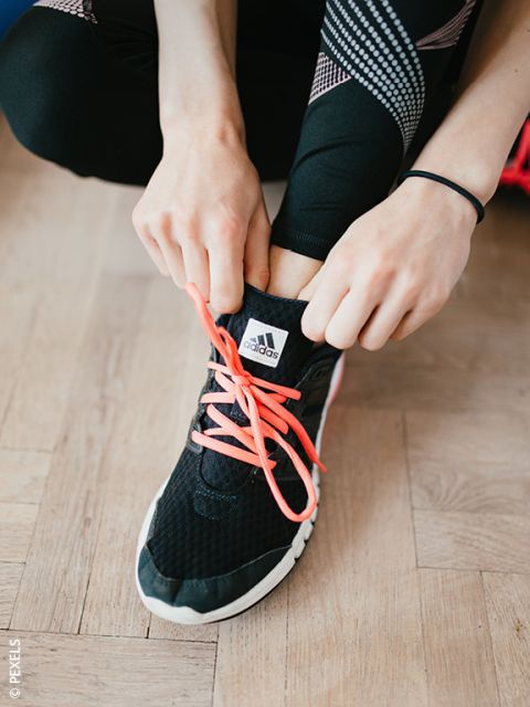 Una persona atando cordones rosa brillante en una zapatilla Adidas negra, mientras lleva leggings negros con estampados blancos y rosas.