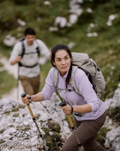 Dos imágenes sobre un fondo de madera muestran a una modelo femenina y a un modelo masculino con ropa de senderismo para exteriores. La modelo femenina lleva un conjunto rosa y verde, el modelo masculino un conjunto verde y sostiene bastones de trekking en la mano. Texto: "TIPO OUTDOOR SENDERISTA ACTIVO".