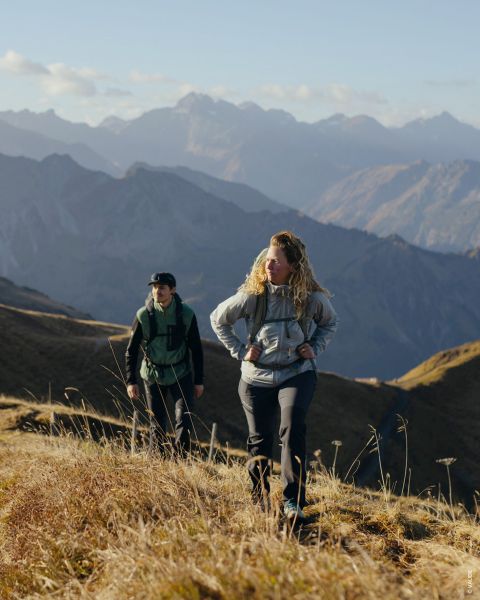 Dos personas se ven en fotos separadas. La foto de la izquierda muestra a una persona con ropa de senderismo y bastones de trekking, la de la derecha a otra persona con mochila y chaqueta. El fondo es una estructura de madera con musgo. El texto dice "Tipo de exterior Senderista de placer".