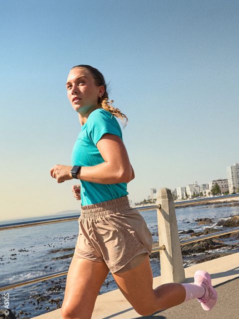 Una mujer con ropa deportiva trota a lo largo del mar en un día soleado, al fondo se ven los edificios de la ciudad.