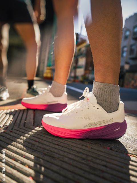 Una persona con zapatillas de correr blancas y rosas está de pie en una acera de la ciudad; la luz del sol hace que los zapatos y las piernas resalten, mientras que el fondo urbano parece borroso.