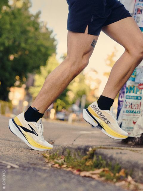 Un corredor en pantalones cortos y zapatillas de correr Brooks amarillas y blancas sube a un bordillo. En el fondo se ven árboles y un poste cubierto de carteles.