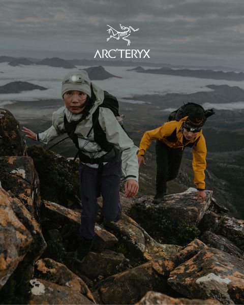 Dos personas caminan por un sendero rocoso de montaña. El cielo está cubierto, y al fondo se ve una cordillera neblinosa. Ambos excursionistas llevan equipo para actividades al aire libre. A la derecha hay un pequeño estanque. Arriba se ve el logo de Arc'teryx.