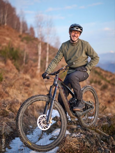 Una persona con casco y chaqueta verde sonríe mientras posa con una bicicleta de montaña en un sendero rocoso y nevado con un fondo de árboles.