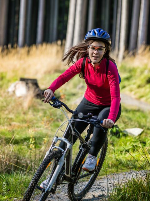 Una mujer con una camiseta roja y un casco monta en bicicleta de montaña por un camino forestal a la luz del sol.