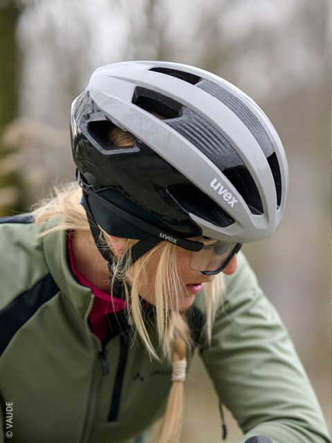 Un hombre con una chaqueta verde y guantes examina un casco de bicicleta negro y está sentado frente a una pared de madera. Lleva una cinta negra para la cabeza con letras blancas.