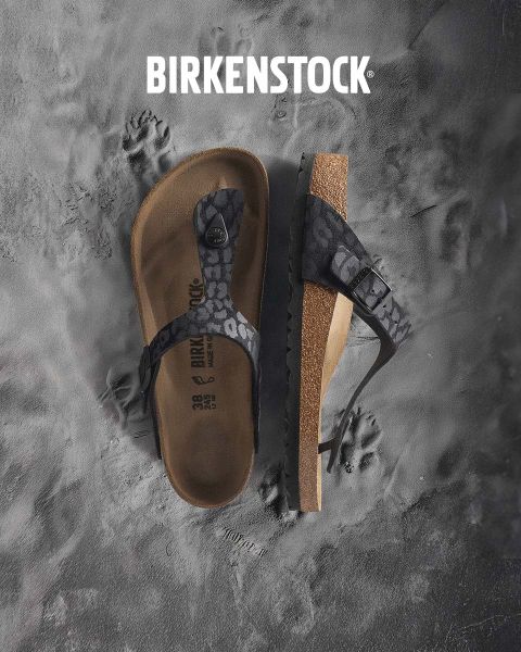 Una persona con sandalias Birkenstock beige está de pie junto a una fuente de agua en un exuberante jardín. El área está rodeada de vegetación y la palabra "BIRKENSTOCK" se ve claramente encima de la imagen.