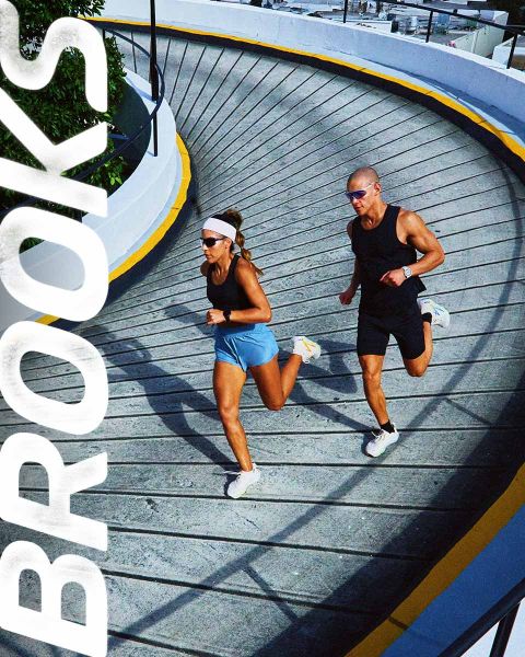 Un grupo de cinco personas corre enérgicamente por un paso de cebra. Llevan ropa deportiva colorida, incluyendo pantalones cortos y camisetas sin mangas. La escena es vibrante, con la palabra "BROOKS" estampada claramente encima de ellos.