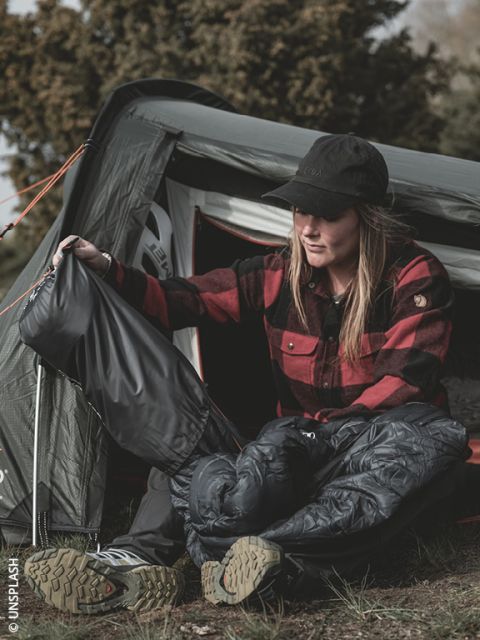 Una mujer con una camisa de cuadros roja está sentada frente a una tienda de campaña, vistiéndose con pantalones de lluvia y botas de montaña, preparándose para actividades al aire libre.