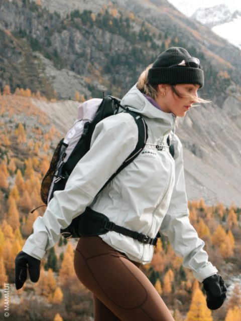 Una mujer camina en la naturaleza en otoño, vestida con una chaqueta, un gorro, guantes y una mochila. Al fondo se ven montañas y naranjos.