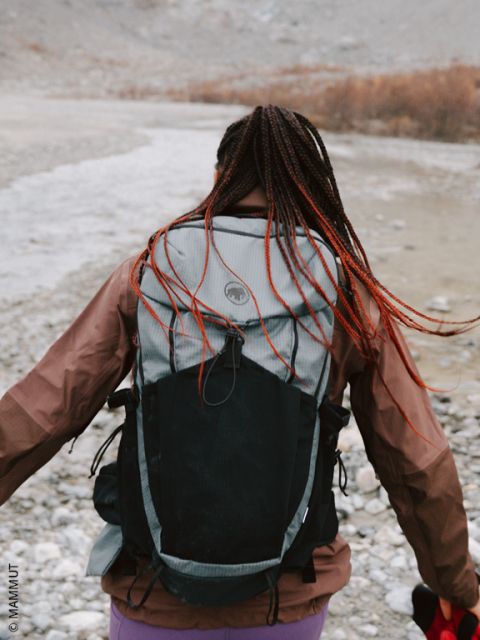 Una persona con el pelo trenzado y una mochila grande camina por un terreno rocoso cerca de un río, de espaldas a la cámara.