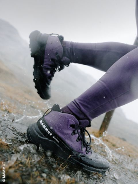 Primer plano de las piernas de una persona con pantalones morados y botas de montaña negras, vadeando un charco en un sendero rocoso y mojado por la lluvia.