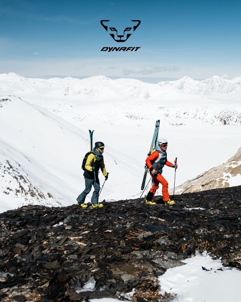 Dos personas corren por un sendero de montaña con una ciudad y un río al fondo. Uno lleva una camiseta negra y pantalones cortos, el otro una camiseta de tirantes azul y pantalones cortos. El logo de Dynafit está en la parte superior de la imagen.