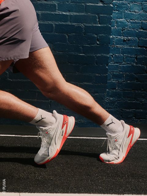 Una mujer con un chándal azul y blanco está sentada en el suelo posando con una rodilla levantada. Lleva calcetines blancos y zapatillas plateadas Under Armour y sonríe levemente frente a un fondo claro y sencillo.