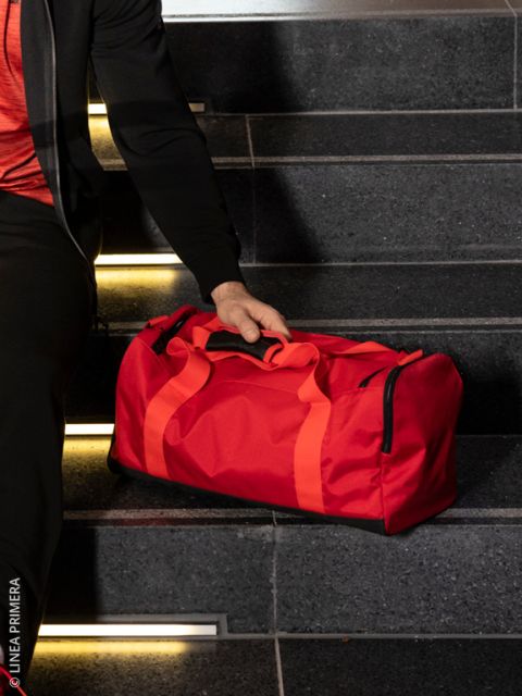 Una persona en pantalones de chándal beige claro, zapatillas negras y top negro sostiene una bolsa de viaje negra brillante. Están de pie sobre tablas de madera oscura junto a un viejo foco y maletas apiladas.